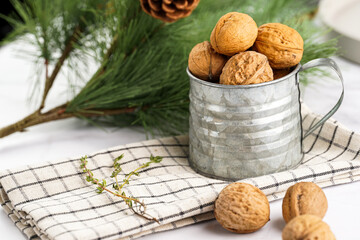 A metal mug with whole brown walnuts on a checkered linen cloth and a christmas fair-tree branch on a white table surface