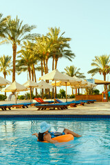 Obraz premium A woman in a sun hat in the pool in a swimming circle with a laptop on the background of palm trees and sun loungers.