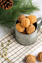 A metal mug with whole brown walnuts on a checkered linen cloth and a christmas fair-tree branch on a white table surface