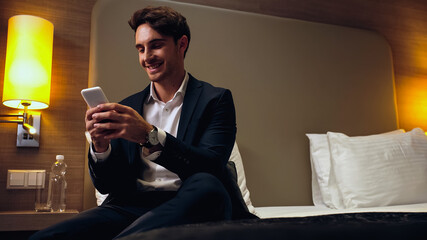 happy businessman sitting on bed and chatting on smartphone in hotel room.