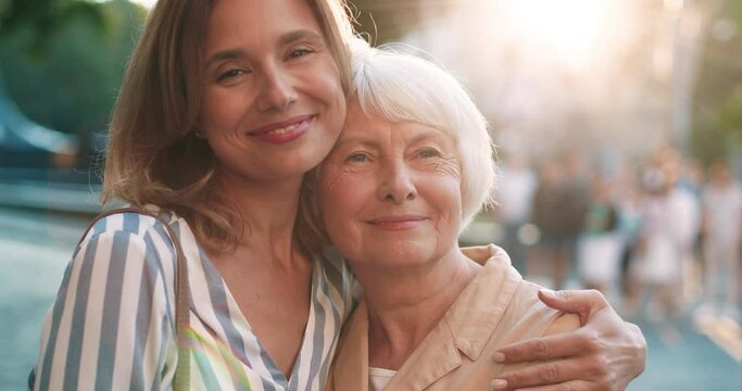 Portrait of happy adult woman and her gray-haired mother standing hugging against the backdrop of street in sunlight and smiling at camera .