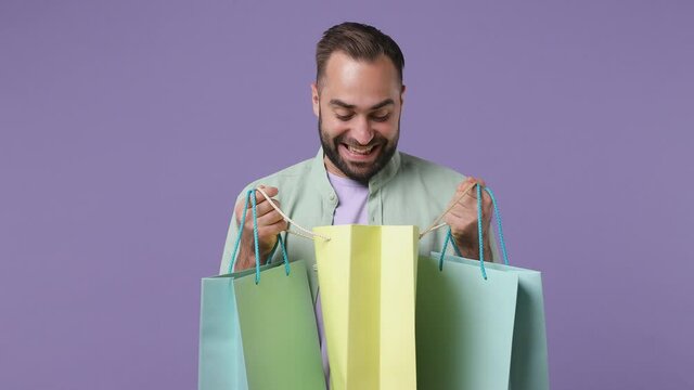 Smiling surprised excited fun young bearded man 20s years old wears mint shirt holding looking into package bags with purchases after shopping isolated on plain light purple background studio portrait