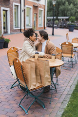happy interracial couple sitting near paper cups and shopping bags in outdoor cafe.