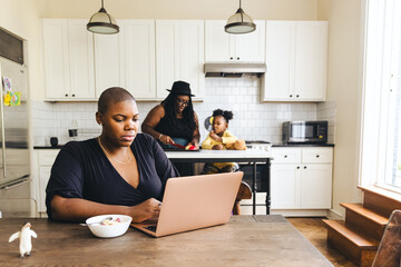 Female freelancer working on laptop while girlfriend and daughter prepare food in kitchen at home