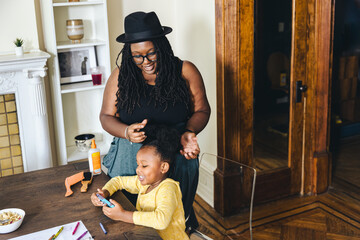 Smiling mother tying hair of daughter using smart phone at home