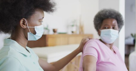 African american female physiotherapist wearing face mask helping senior female patient exercise - Powered by Adobe