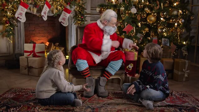 Santa Claus gives gifts to two children: a boy and a girl. The mood is one of joy and magic