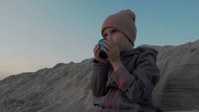 Closeup of little child girl in warm clothes at cold weather having a hot beverage at sandy coast during sunset 