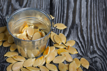 Metal bucket with pumpkin seeds. A few seeds are scattered around. On black pine boards. Close-up shot.