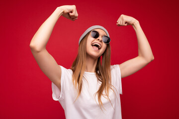 Portrait of young emotional positive happy attractive dark blonde woman with sincere emotions wearing casual white t-shirt with empty space for mockup grey hat and sunglasses isolated over red