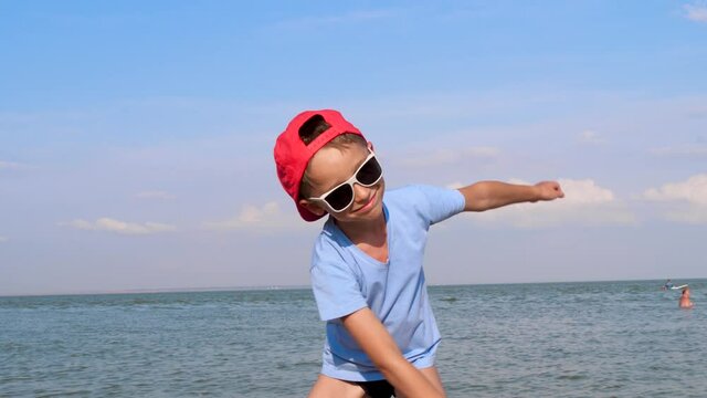 A cheerful child in sunglasses and a baseball cap is dancing on the beach. An active child. Imagination and a dream.