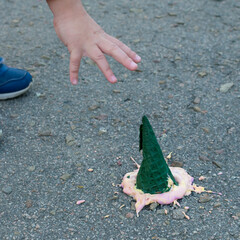 An upset child's hand reaches for the ice cream that has fallen on the pavement. Bright ice cream in a green cone fell on the asphalt.