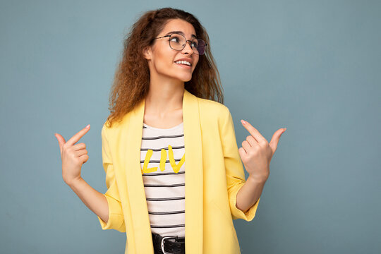 Photo Of Young Positive Beautiful Brunette Curly Woman With Sincere Emotions Wearing Casual Yellow Jacket And Optical Glasses Isolated On Blue Background With Copy Space And Pointing At Yourself