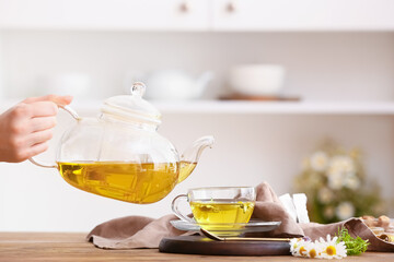 Woman pouring fresh chamomile tea from teapot into cup on wooden table