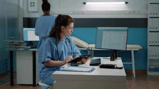 Medical assistant typing on tablet screen and checking computer for appointment in doctors office. Nurse looking at monitor while using modern gadget for healthcare checkup visit.