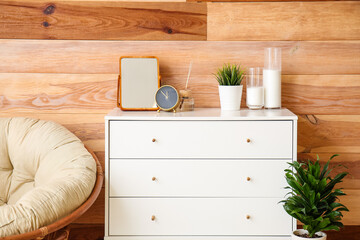 Golden alarm clock on stylish chest of drawers with decor near wooden wall