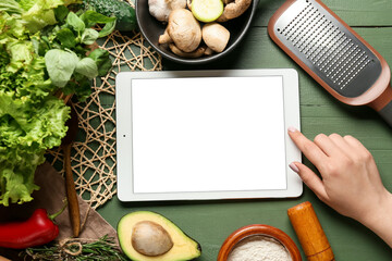Woman using digital recipe book, different ingredients and kitchen utensils on green wooden background