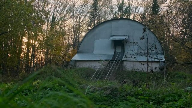 A wierd hangar or old shed with the semi-sphere roof and a metal ladder seems to be in the middle of nowhere. Industrial premises somewhere in a factory area among usual trees and coniferous trees.