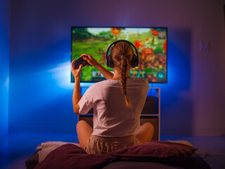A blond girl with headphones and a joystick in her hands sits in her bedroom in front of a large...
