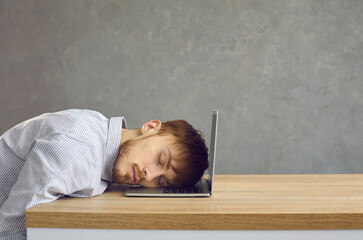 Tired male office worker sleeping sitting at a desk putting his head on a laptop keyboard. Side view of a young man on a gray background with space for text. Concept of burnout, fatigue and laziness.