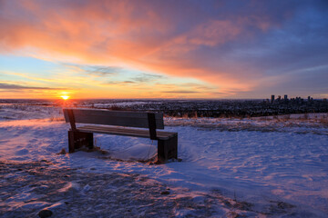 Wide angle view of the sun rising over a city with an empty bench in the foreground
