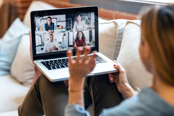  Back view of mature business woman speaking on video call with diverse colleagues on online briefing with laptop on sofa at home.