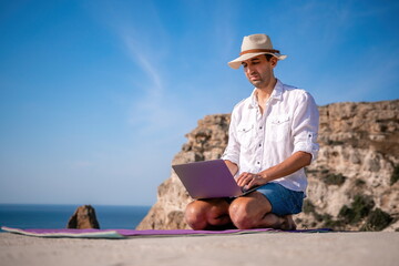 Fototapeta premium A digital nomad, a man in a hat, a businessman with a laptop does yoga on the rocks by the sea at sunset time, does a business operation online from a distance. Remote work on vacation.