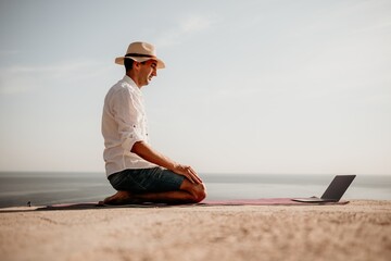 A digital nomad, a man in a hat, a businessman with a laptop does yoga on the rocks by the sea at sunset time, does a business operation online from a distance. Remote work on vacation.