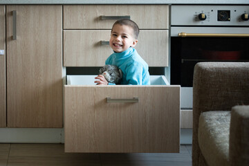Preschool boy sitting in kitchen drawer with his toy and smiling at home 