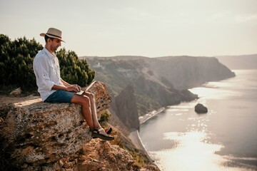 Digital nomad, man in the hat, a businessman with a laptop sits on the rocks by the sea during sunset, makes a business transaction online from a distance. Remote work on vacation.