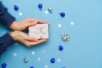Woman's hands holding gift box present, tied with ribbon bow on blue background around christmas...
