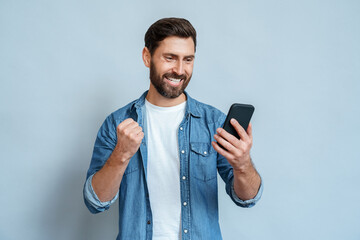 Happy delighted satisfied adult man looking at mobile phone screen gesturing yes winning pose,...