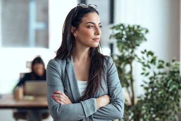 Beautiful young woman thinking while waiting looking forward standing next to the window at the office.