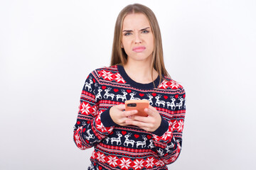 Portrait of a confused Young beautiful caucasian girl wearing christmas sweaters on white background holding mobile phone and shrugging shoulders and frowning face.