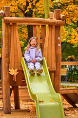 The girl went down the hill. The child plays on the playground in the fall. Wooden platform on the territory of the new house. Happy childhood. Yellow foliage on the trees on a sunny day.