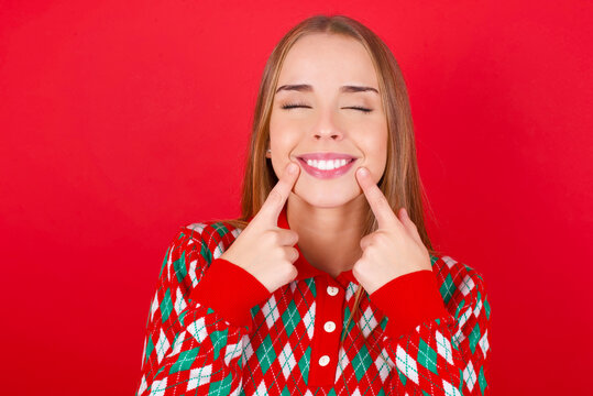 Happy Young beautiful caucasian girl wearing christmas sweaters on red background with toothy smile, keeps index fingers near mouth, fingers pointing and forcing cheerful smile