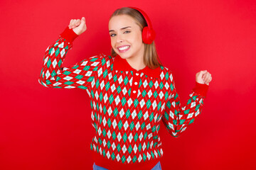 Carefree Young beautiful caucasian girl wearing christmas sweaters on red background with toothy smile raises arms dances carefree moves with rhythm of music listens music from playlist via headphones