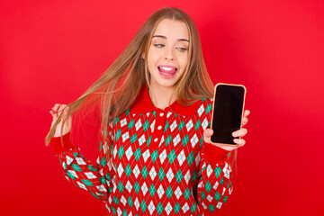 Photo of nice pretty Young beautiful caucasian girl wearing christmas sweaters on red background demonstrate phone screen hold hair tails