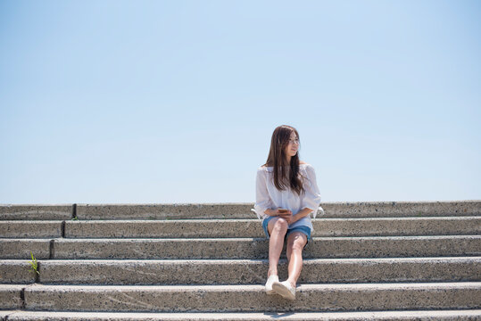 海の堤防にスマホを持って座る女性