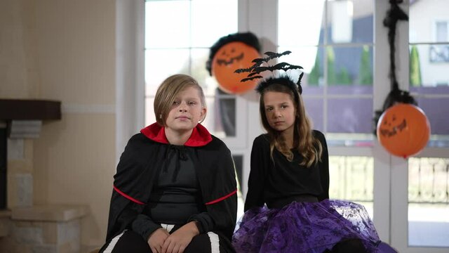 Caucasian boy and girl in Halloween costumes sitting in living room watching TV as woman alien walking behind glass door at background. October 31 holiday celebration and traditions. Slow motion