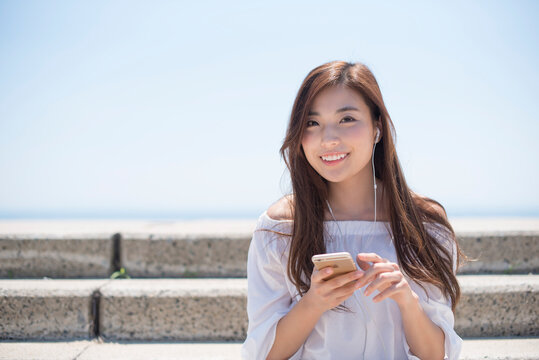 海の堤防に座ってスマホで音楽を聴く女性