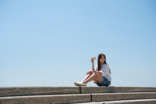 海の堤防に座ってスマホで音楽を聴く女性