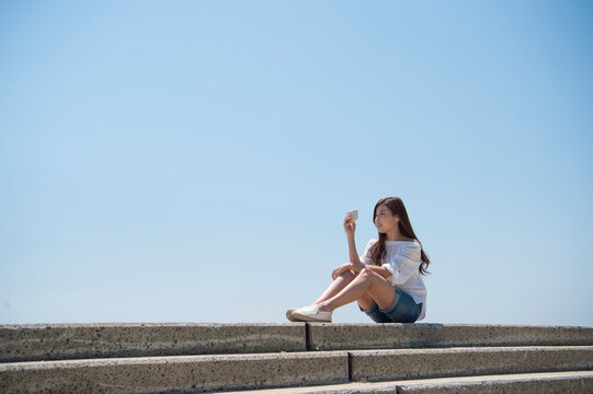 海の堤防に座ってスマホで音楽を聴く女性