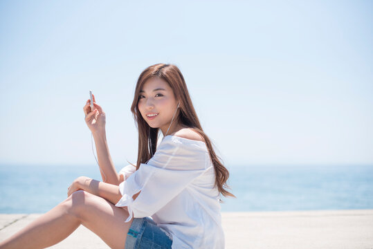 海の堤防に座ってスマホで音楽を聴く女性