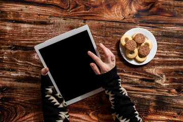 Woman's hands holding digital tablet on the wooden table with a plate full of cookies