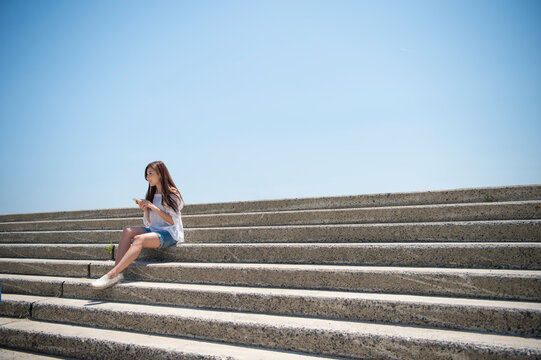 海の堤防に座ってスマホで音楽を聴く女性