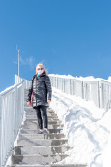 An adult woman wearing a coat, sunglasses and a face mask standing on step of a staircase with a lot of snow around her