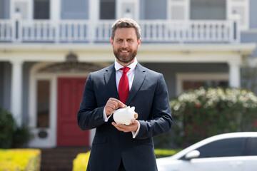Fototapeta na wymiar Happy young man putting money in piggy bank. Businessman holding piggybank on rich house background. Save money and financial investment.