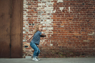 Fototapeta na wymiar Little cute girl in a blue t-shirt and jeans with a cap is dancing on the background of an old brick wall.