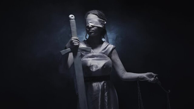 Greek goddess Themis blindfolded holds sword and raise scales, turning head on black background with smoke. Woman with eyes closed hold sword in hand and raise scale. Studio. Concept of legal justice